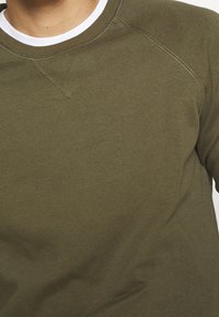 Uomo che indossa un maglione girocollo verde oliva sopra una t-shirt bianca, mostrando la parte superiore del petto e l'area del collo.