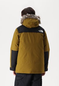 Żółto-czarna kurtka z kapturem, wykończona sztucznym futrem u kołnierza, z logo na lewym ramieniu i gładkim, teksturowanym wykończeniem.