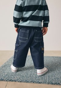 Pantalones cargo de mezclilla, azul oscuro con costuras blancas, presentan múltiples bolsillos y un corte relajado. Combinados con zapatillas blancas sobre una alfombra texturizada.