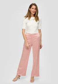 Witte top met korte mouwen en een unieke halslijn, gecombineerd met lichtroze wijde broek. Model draagt beige sandaaltjes met banden.