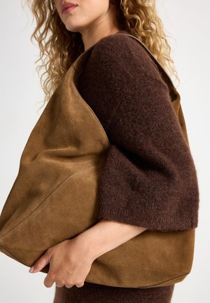 Borsa tote in suede marrone con una texture morbida, grande forma e spallaccio largo, tenuta in una mano accanto a un maglione peloso marrone scuro.