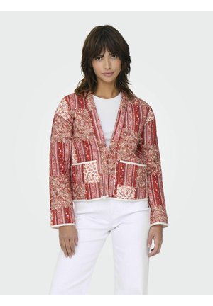 Femme portant une veste rouge à motifs floraux et rayés, un t-shirt blanc et un pantalon blanc, debout devant un fond uni.