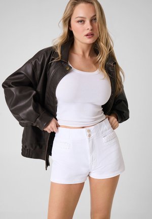 Femme blonde portant une veste en cuir marron foncé sur un débardeur blanc et un short blanc taille haute, posant devant un fond uni.