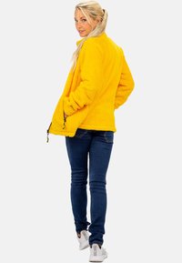 Geel fleece jack met een textuurovervlak, ritszakken en een ontspannen pasvorm, gecombineerd met donkere jeans en witte sneakers.