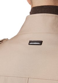 Beżowa odzież wierzchnia o teksturowanej powierzchni, z brązowym prążkowanym kołnierzem i prostokątną czarną metką w okolicy dekoltu.