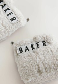 Jasnoszare, puszyste kapcie z materiałową etykietą z napisem „BAKER” w czerni na białym tle. Zaokrąglony kształt z miękkiego materiału.