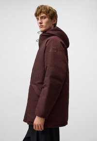 Parka isolante bordeaux con cappuccio, tessuto testurizzato, tasche laterali e logo ricamato sulla manica. Design pulito e moderno.