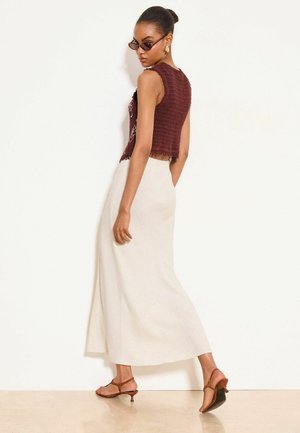 Kobieta w brązowym, bezrękawnikowym, szydełkowym topie, kremowej długiej spódnicy, brązowych butach na niskim obcasie, okularach przeciwsłonecznych i złotych kolczykach-kołach, pozująca bokiem.