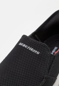 Svart slip-on sko laget av teksturert mesh-materiale, med en hvit "SKECHERS"-logo på overdelen. Elastiske sidepaneler for enkel påkledning.
