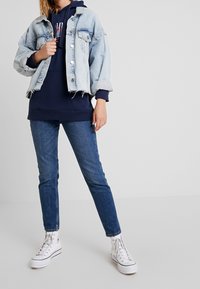 Jeansjacka med en ljusblå tvätt, uppvikta ärmar och fransad nederkant. Under jackan en mörk marinblå hoodie. Tätt sittande blå jeans och vita högre sneakers.