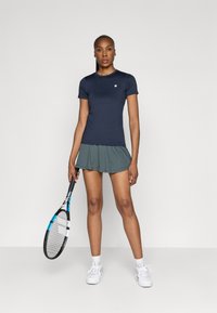 Tengerészkék, rövid ujjú sportpóló szürke, bővülő teniszkabáttal párosítva. A modell egy teniszütőt tart a kezében, fehér sportcipőt visel.