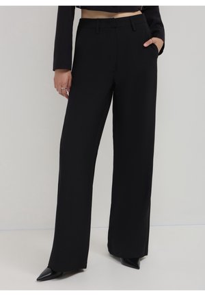 Femme portant un pantalon noir taille haute à jambes larges avec des poches latérales et des chaussures noires à bouts pointus sur un fond uni.