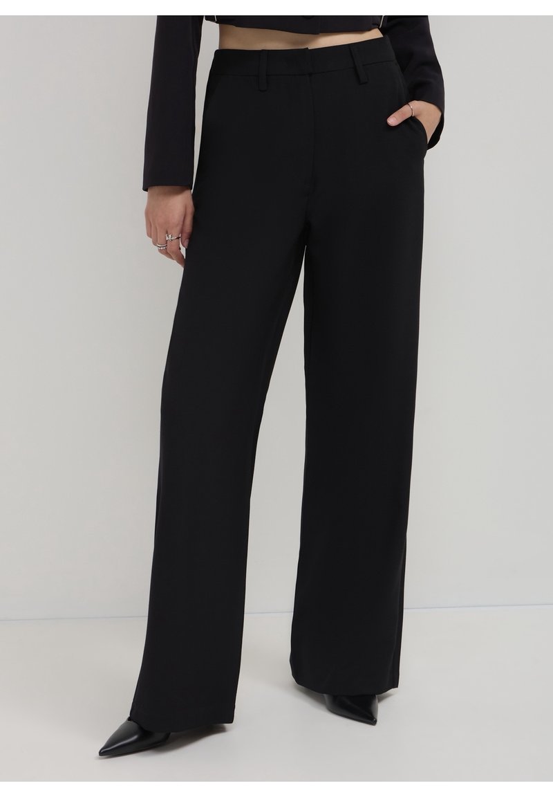 Femme portant un pantalon noir taille haute à jambes larges avec des poches latérales et des chaussures noires à bouts pointus sur un fond uni.