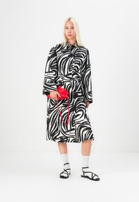 Rochie cămașă cu model zebrat alb-negru, dotată cu un cordon, asortată cu o geantă roșie, șosete albe și sandale negre. Textură de țesătură netedă.