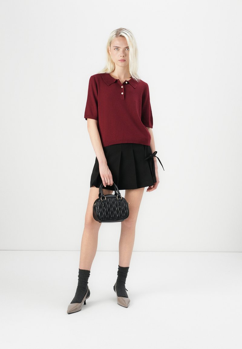 Bordowa dzianinowa koszulka polo z kołnierzykiem, czarna plisowana spódnica, czarna torebka o teksturowanej powierzchni, szare skarpetki oraz dwukolorowe czółenka w szpic.