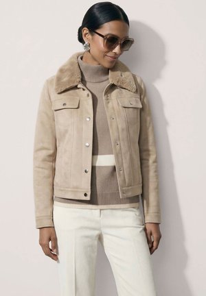 Frau trägt beige Wildlederjacke mit Fellkragen, beige gestreiften Rollkragenpullover, weiße Hose, große Ohrringe und übergroße Sonnenbrille.