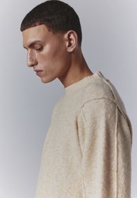 Pull en tricot crème avec une finition texturée, un col en côtes et des manches raglan, présenté sur un fond gris doux.