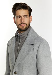 Grau wohlschmeckender Mantel mit einem doppelten Verschluss, großen Revers und einem auffälligen Knopf. Darunter eine graue isolierte Jacke mit hohem Kragen.