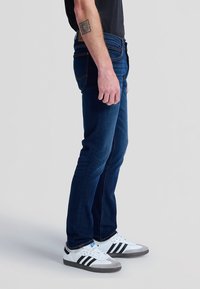 Homme portant un jean bleu foncé, une chemise noire, des baskets blanches Adidas avec des rayures noires et un tatouage carré sur l'avant-bras gauche, debout de côté.