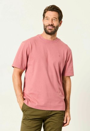 Bearded man with short brown hair wearing a faded rose short-sleeve t-shirt and olive green pants, standing with one hand in pocket.