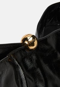 Bolso de mano de cuero negro con un acabado brillante, de textura suave y un destacado cierre circular dorado que muestra el nombre de la marca en relieve.