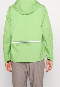 Veste légère verte avec une finition texturée, une capuche et un accent de fermeture éclair réfléchissante sur le dos. Pantalon gris visible en dessous.