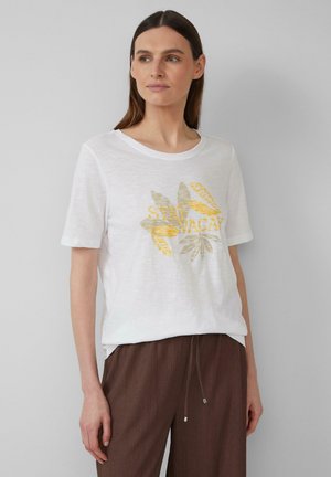 T-shirt blanc à manches courtes en tissu léger, arborant un graphique de feuilles dorées et vertes avec le texte "STAY VACAY". Associé à un pantalon texturé marron.