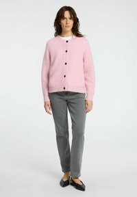 Gilet en laine rose à col rond, doté de cinq boutons noirs, associé à un jean droit gris et des ballerines noires. Texture lisse.