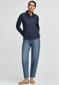Granatowa koszula zapinana na guziki, wykonana z gładkiego materiału, z długimi rękawami, klasycznym kołnierzykiem i małym czerwonym logo na piersi. Połączona z niebieskimi dżinsami.