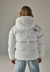 Doudoune blanche avec capuche, design matelassé, coutures visibles et logo au dos. Associée à un jean bleu clair.
