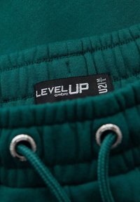 Cintura de chándal en color verde azulado con elasticidad y cordón. Presenta un parche de tela negra con el logo "LEVEL UP" y detalles de cuidado. Textura suave.