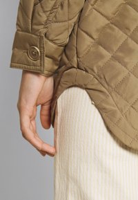 Gequilted bruine jas met een knoopsluiting aan de manchetten, gecombineerd met lichte gestreepte broeken. De stof lijkt glad met een zachte textuur.