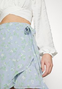 Světle modrá květinová zavinovací sukně vyrobená z lehkého materiálu, s jemnými zelenými a bílými vzory, uvázaná na boku mašlí.