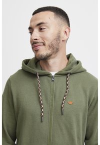 Sudadera verde con cremallera y capucha ajustable, que presenta acentos trenzados y un pequeño logo marrón en el pecho. Textura de tela suave.