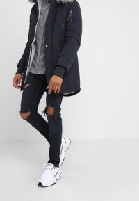 Person med marinblå parka med pälsmössa, grå skjorta med knappar, trasiga svarta skinny jeans och vita Nike-sneakers framför en neutral bakgrund.
