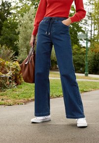Pantalon en jean bleu foncé à jambes larges avec taille à cordon et poches latérales, porté avec des baskets blanches. Sac à main marron tenu dans une main.