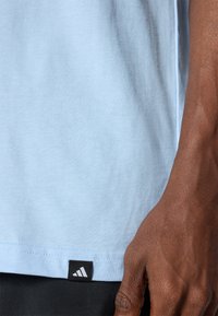 Šviesiai mėlynas medvilninis marškinėliai su apvaliu kaklu, kurių apačioje yra juodas Adidas logotipas, pagaminti iš lygaus, šiek tiek tekstūruoto audinio.