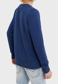 Felpa blu navy realizzata in morbido cotone, con colletto rotondo, polsini e orlo a costine. Indossata sopra jeans chiari strappati.