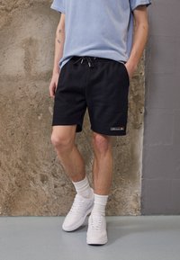 Shorts noirs avec taille élastique et cordon de serrage, tissu texturé, poches latérales et logo brodé. Assortis à des baskets blanches et des chaussettes.