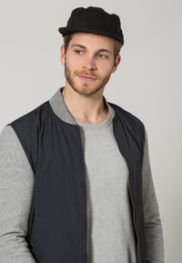 Mustaga kork, hall pikka varrukaga särk ja must bomber-stiilis vest ribakraega. Omab lukuga esist ja küljetaskuid.