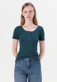Teal pruhované tričko s krátkým rukávem, které má kulatý výstřih a tříknoflíkovou légu na hrudi. V kombinaci s modrými džíny s vysokým pasem.