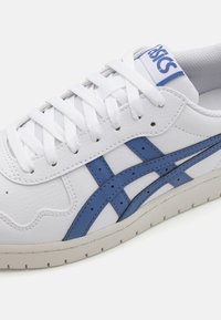 Baltie sneaker ar ziliem akcentiem un logo; tam ir teksturēta ādas augšdaļa, apaļš purngalis un plakani baltie šņorēšanas diegi virs mīkstinātās mēles.