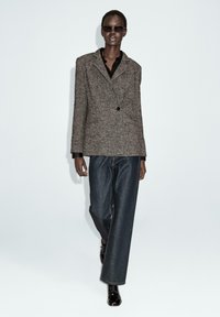 Blazer marrone a spina di pesce con chiusura a un bottone, abbinato a jeans in denim scuri a gamba larga. Il modello è in piedi contro uno sfondo chiaro.