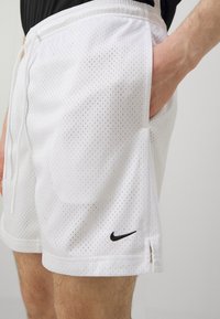 Balti perforuoti sportiniai šortai su elastinga juosta, raišteliu, šoninėmis kišenėmis ir juodu Nike logotipu kairėje kojoje.