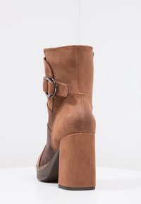 Bottine en daim marron avec un talon épais, dotée d'une boucle décorative et d'une texture lisse. Le design latéral présente un panneau en matériau contrastant.