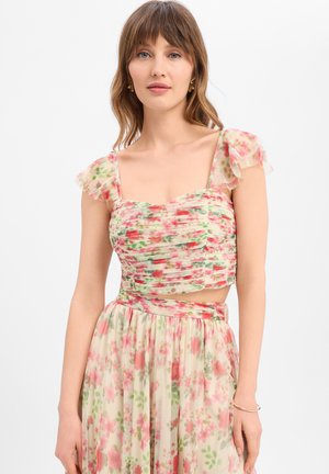 Junge Frau trägt ein florales, rosa-grünes transparentes Crop-Top mit Rüschenärmeln und passendem hoch tailliertem Rock.
