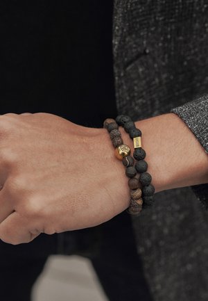 Två beadade armband på ett handled: det ena med matta svarta lavastenar och ett guldaccent, det andra med texturerade bruna och svarta pärlor.