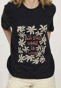 Personne portant une chemise noire avec des fleurs crème et le texte "Tout ce dont vous avez besoin, c'est de l'amour" en lettres orange-brun sur le devant.