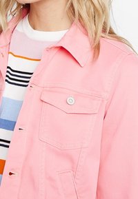 Chaqueta de mezclilla rosa con bolsillos abotonados, cuello y costuras visibles. Usada sobre una camiseta de rayas horizontales multicolores.