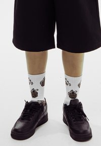 Zapatillas negras de Nike, calcetines blancos con estampado de piñas marrones y pantalones cortos negros hasta la rodilla. Textura suave y diseño casual.